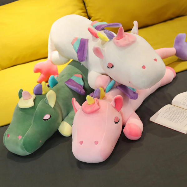 3 grandi peluche di unicorno, i due in basso sono verdi e rosa e quello in alto è bianco, può vedere un divano giallo sullo sfondo e sono posizionati su un pavimento nero