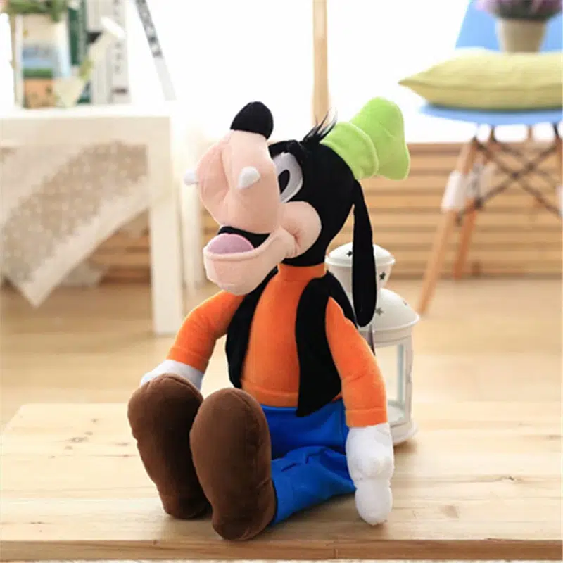 Il personaggio Disney di peluche, seduto su un pavimento di legno, indossa pantaloni blu e un top arancione