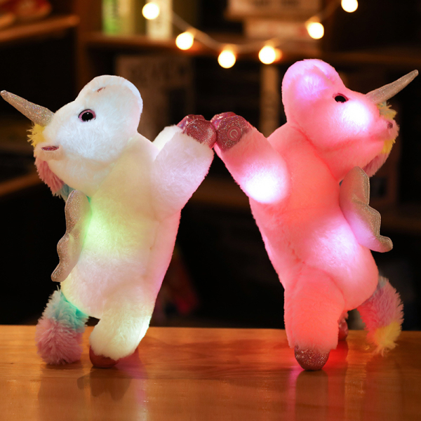 Su un tavolo di legno, 2 peluche unicorno, uno bianco e uno rosa, sono sollevati sulle gambe posteriori e si tengono per quella anteriore e i loro corpi sono illuminati