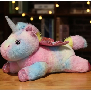 Su un tavolo di legno un peluche di unicorno multicolore è sdraiato a pancia in giù con scaffali e luci sullo sfondo